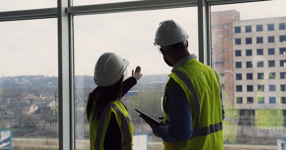 Two,construction,engineers,standing,indoors,near,big,window,looking,at